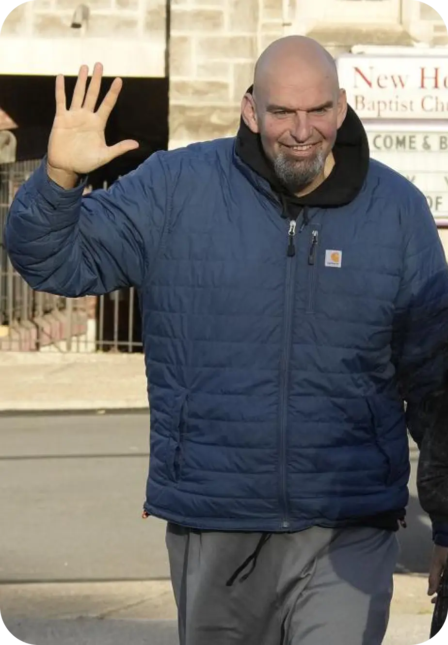 A bald man with a beard in a blue jacket has his right hand up waving.