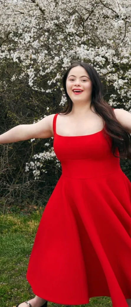 A person standing in front of a blooming tree in a red dress with their arms outstretched.