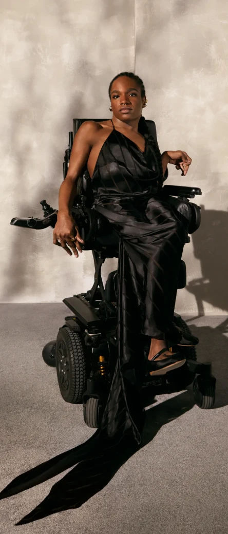 A person sitting in a wheelchair with a black dress on. The dress is pleated at the top and has a long train shown draping in front of the wheelchair.